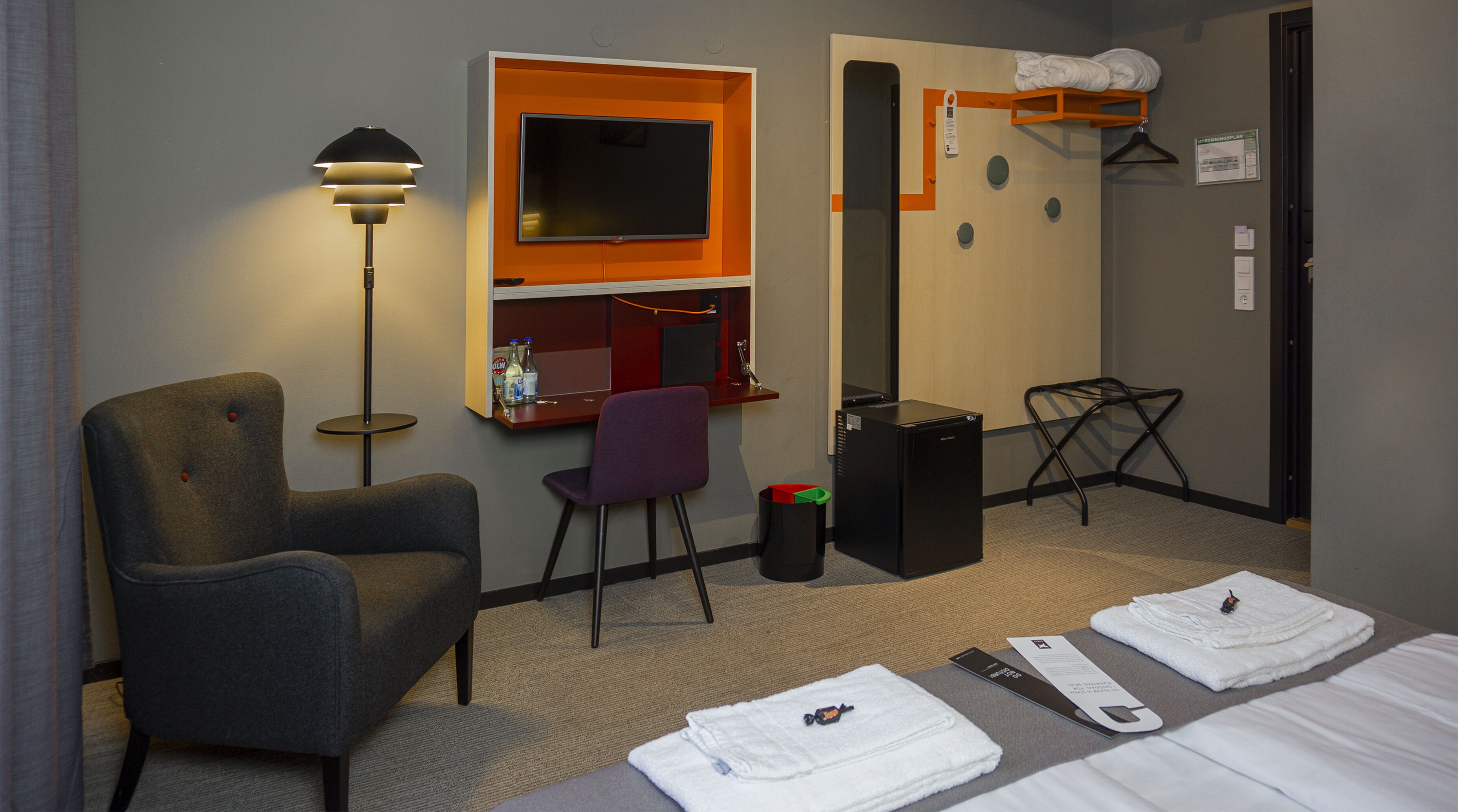 A hotel room with a bed, a desk with a chair, a TV, a lamp, a trash bin, and a coat rack with a towel.