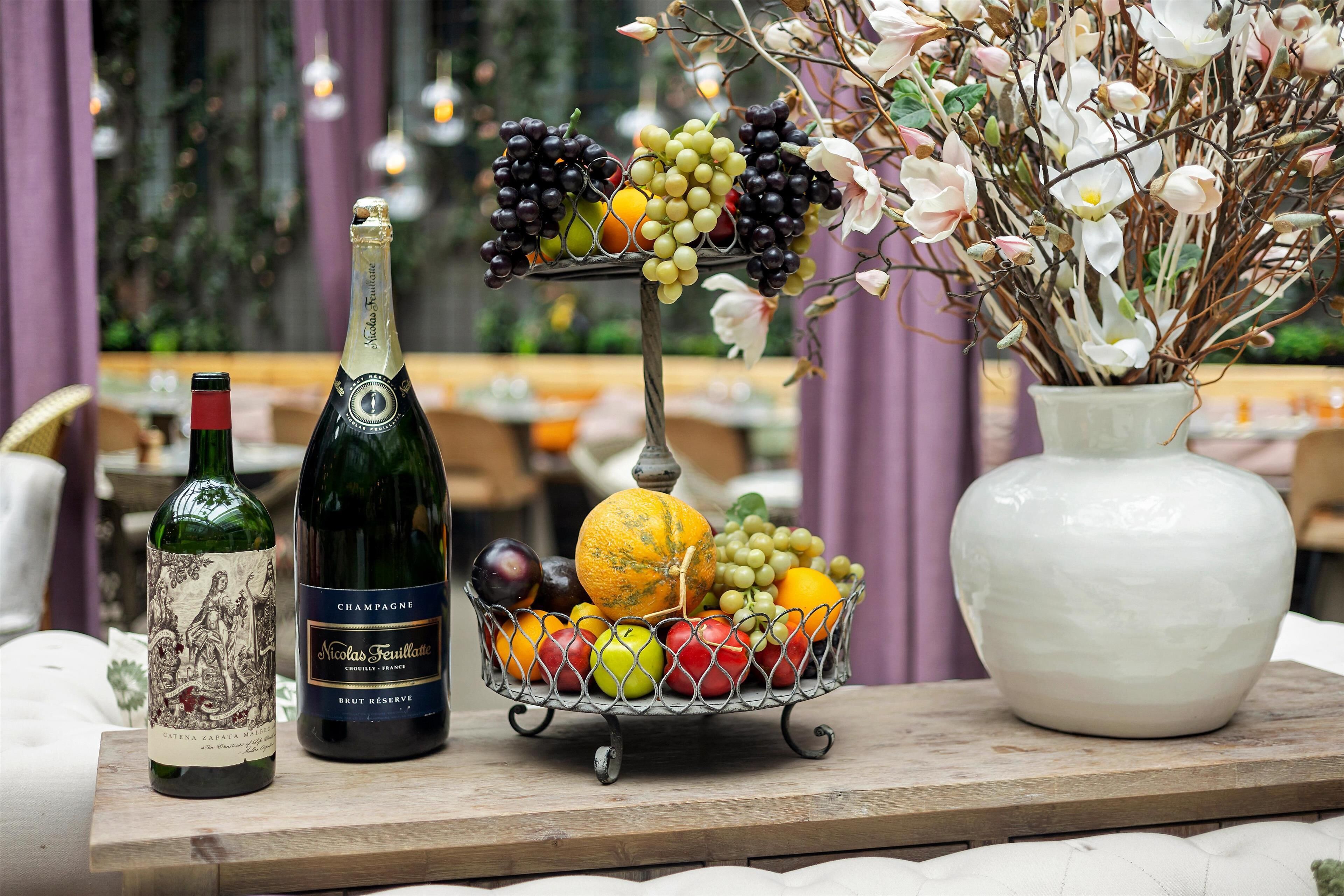 Een tafel met een fles champagne, een fruitmand en een bloemenvaas.