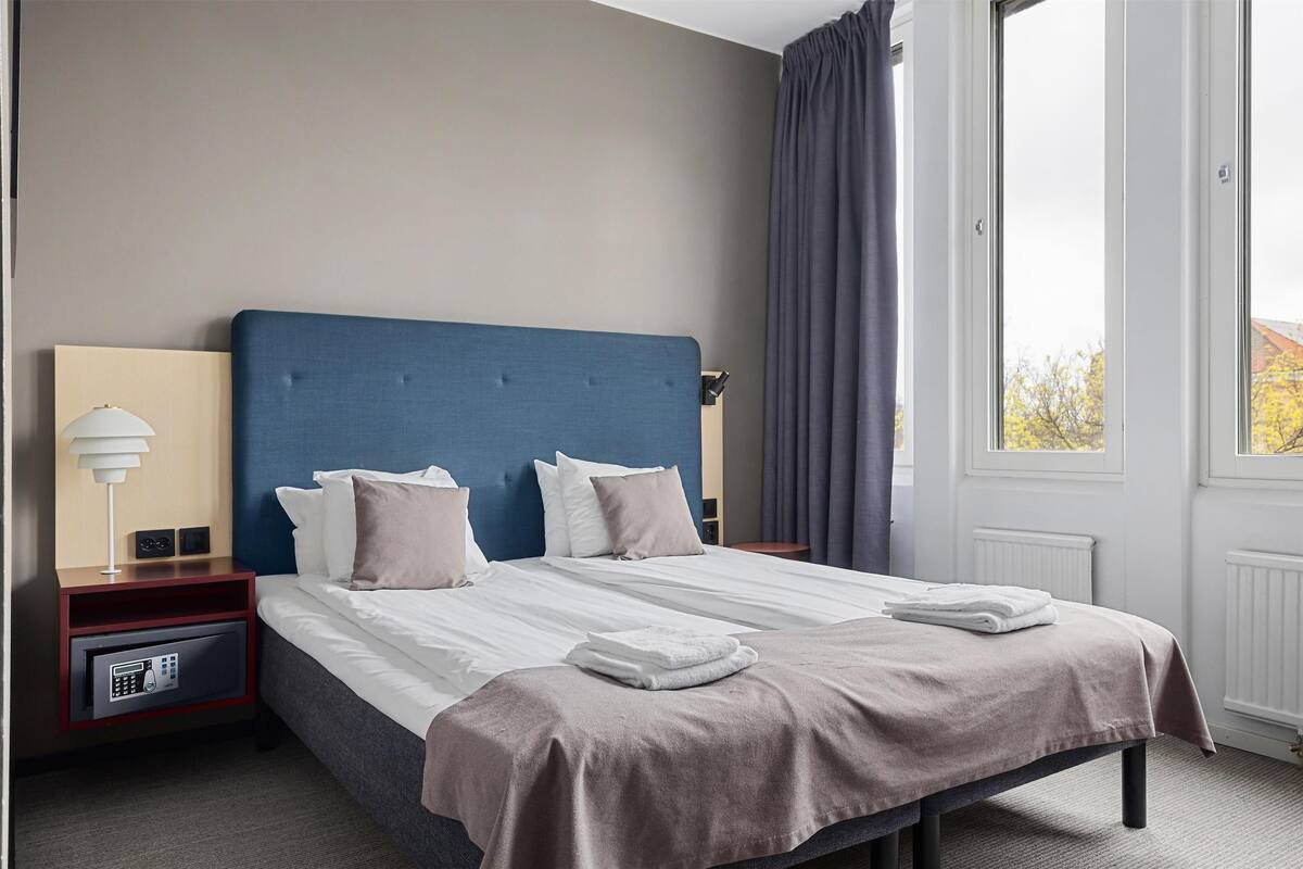 A hotel room with a large bed, a nightstand with a lamp, a safe, and a view of the outside through a window.