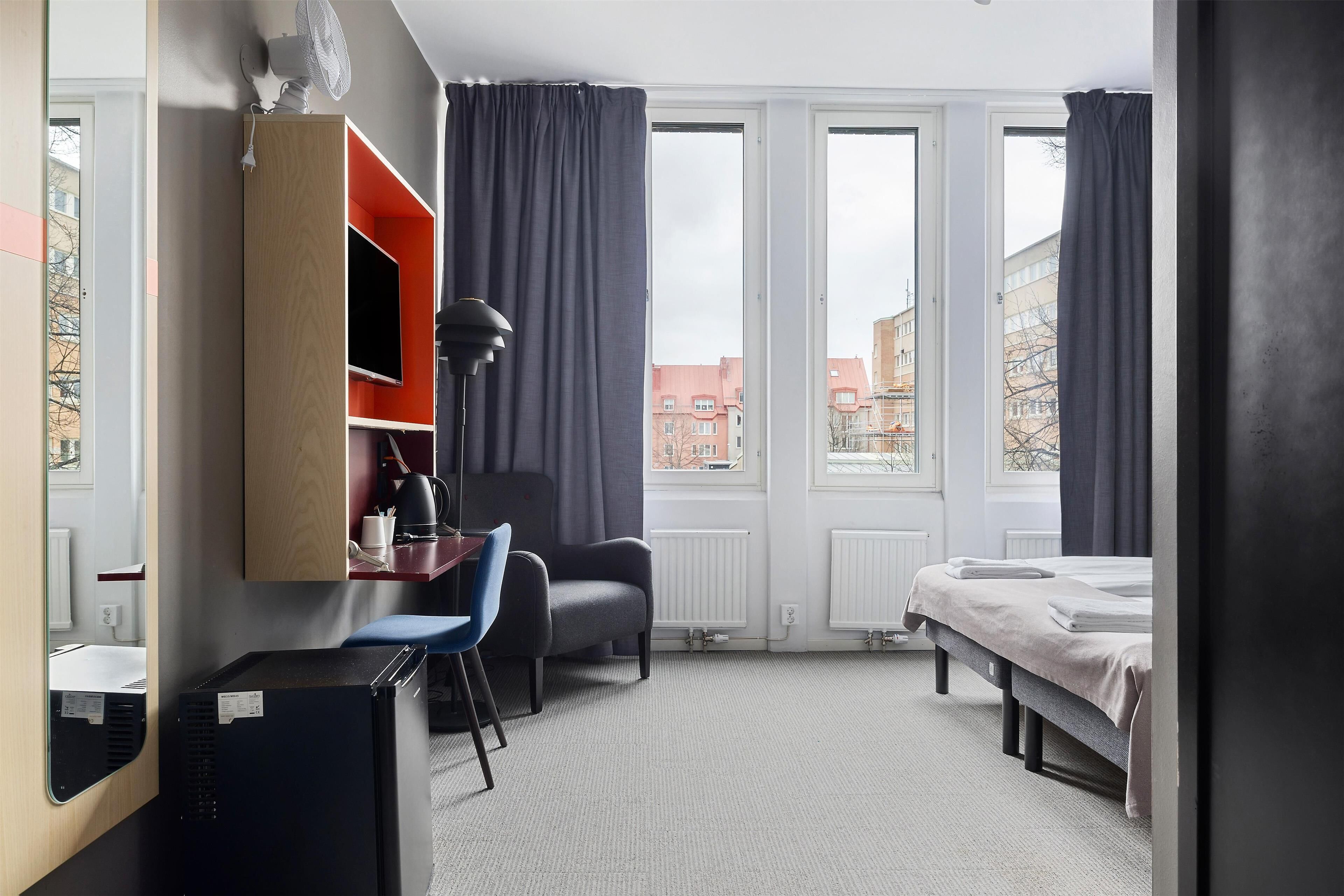 Habitación de hotel con cama, escritorio, silla, televisor y ventana con cortinas.