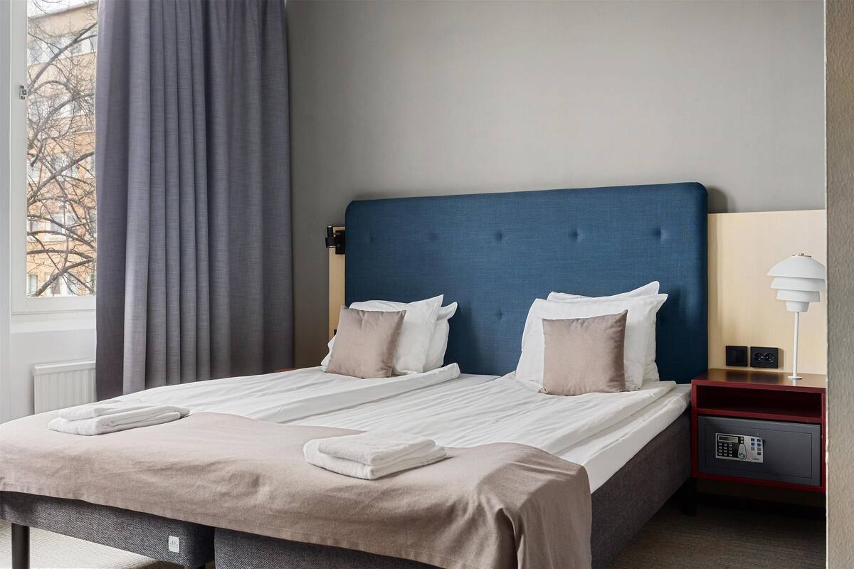 A hotel room with a large bed, a nightstand with a lamp, and a view of trees outside.