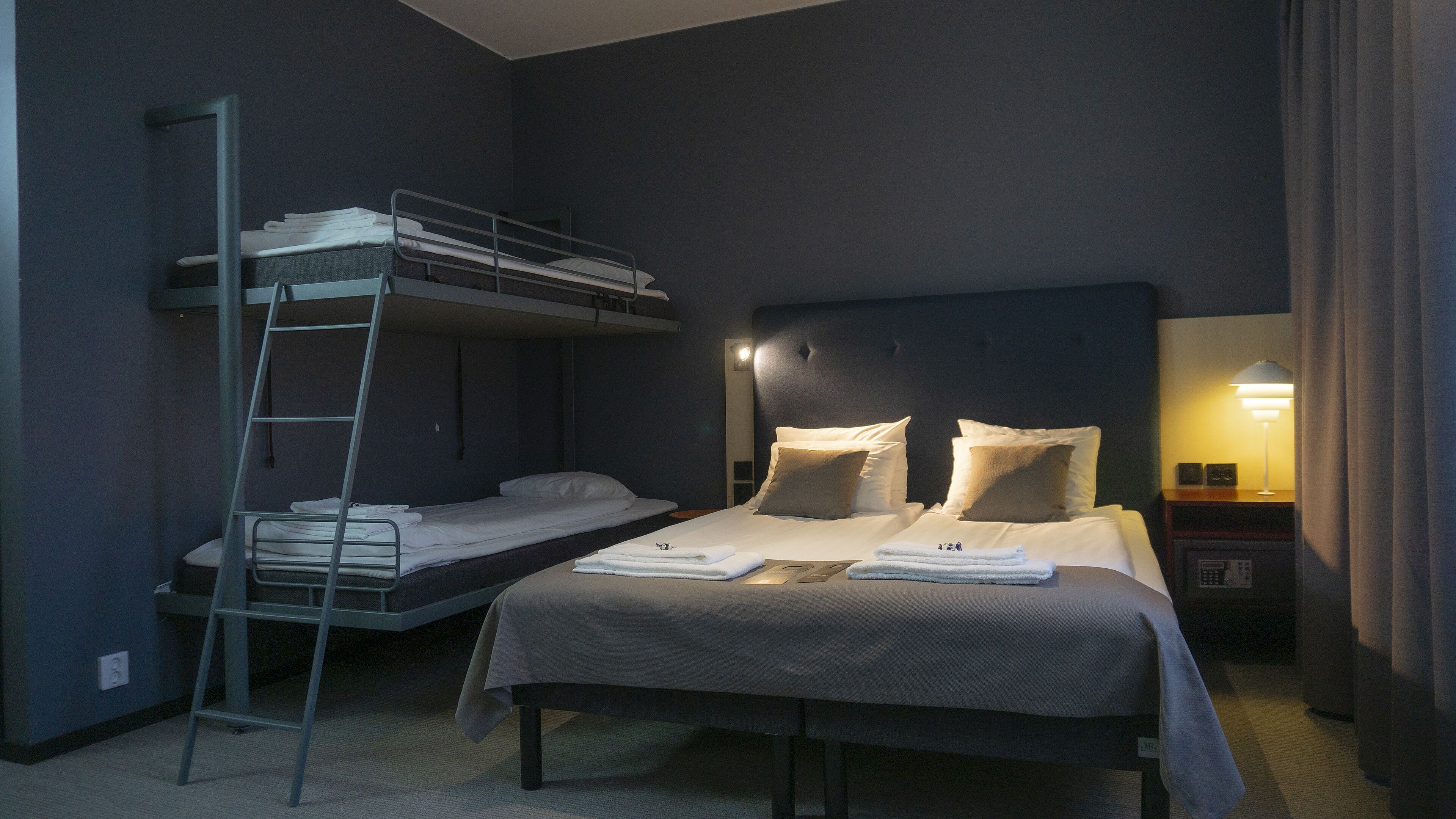 Ein Hotelzimmer mit Etagenbett, einem Einzelbett, einem Nachttisch mit Lampe und einem an der Wand montierten Fernseher.