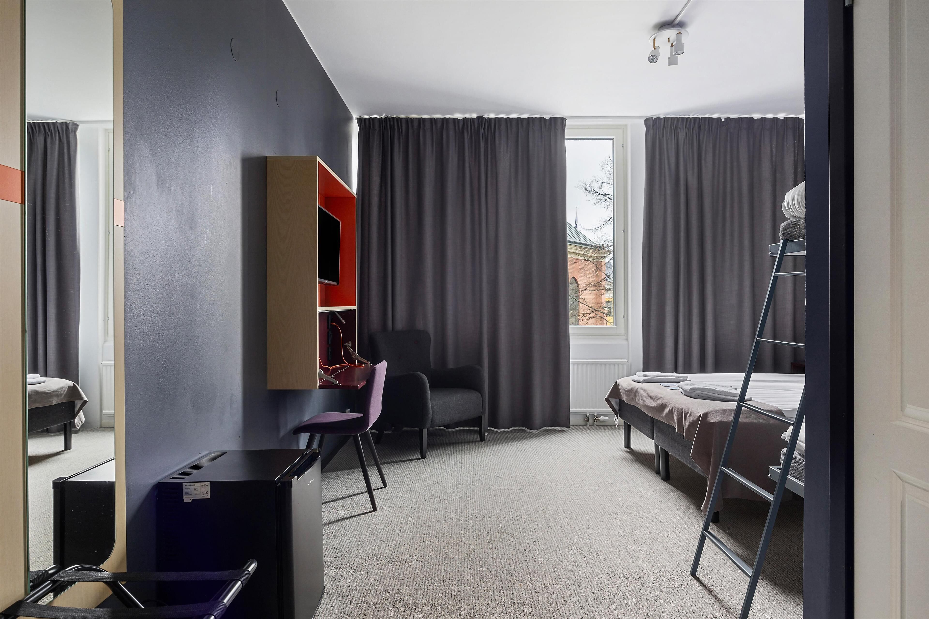 Habitación de hotel pequeña con cama, escritorio, silla, armario y ventana con cortinas.