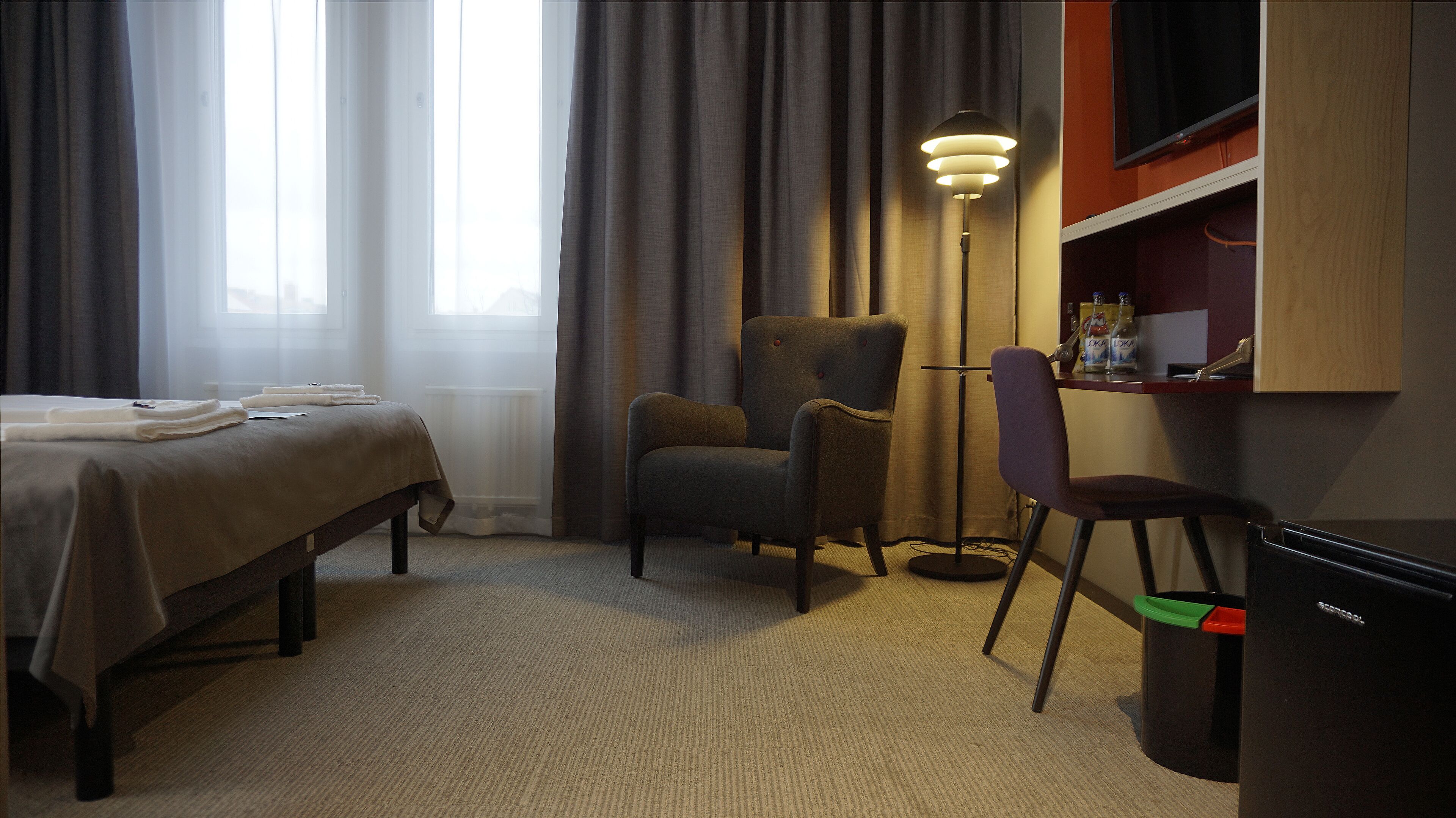 A hotel room with a bed, two armchairs, a desk with a lamp, and a trash bin.