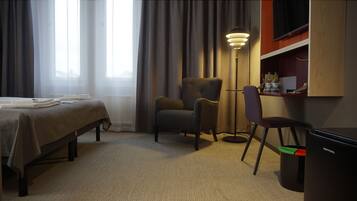 A hotel room with a bed, two armchairs, a desk with a lamp, and a trash bin.