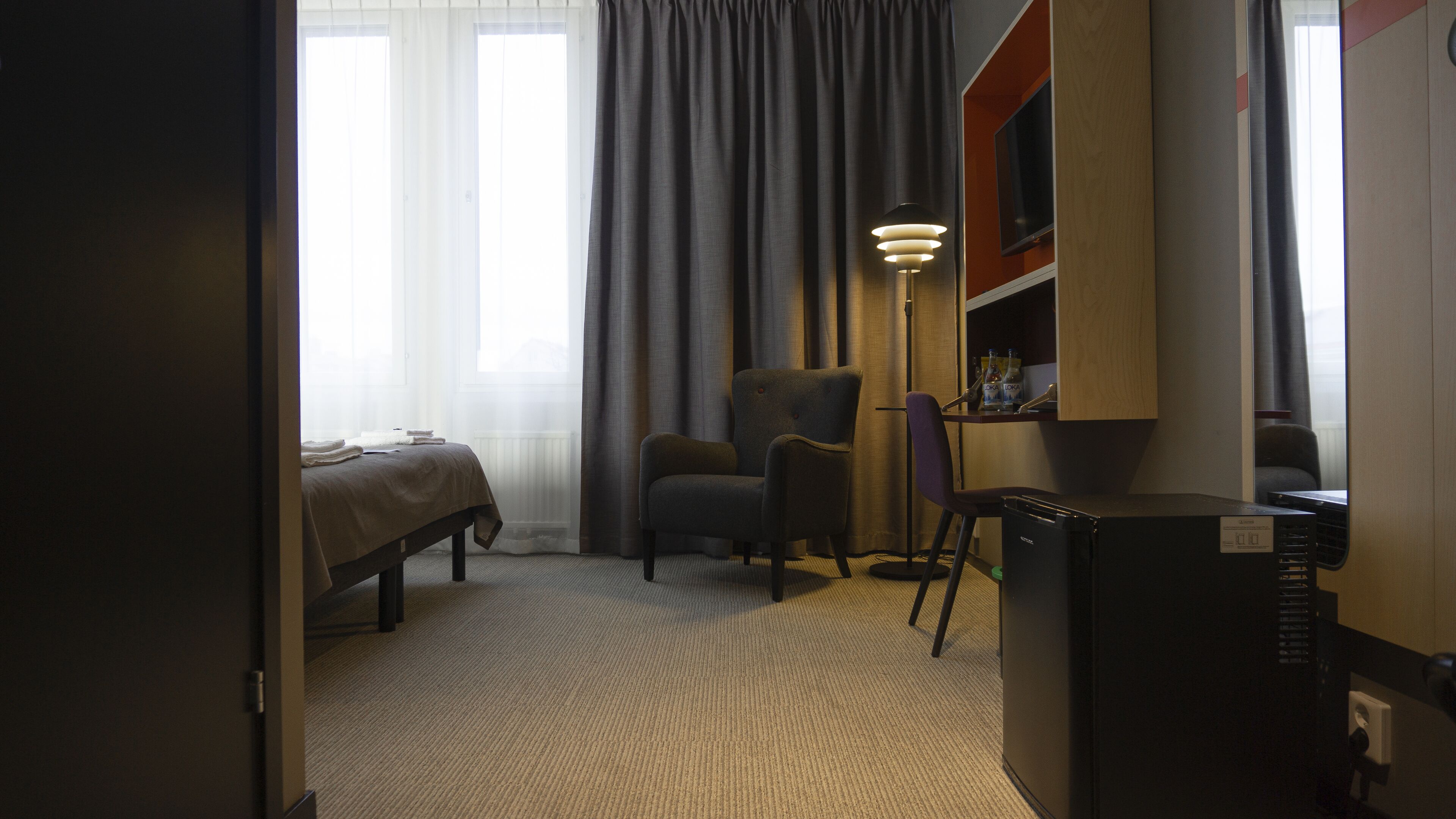 A hotel room with a bed, a chair, a desk with a computer, a lamp, and a TV.