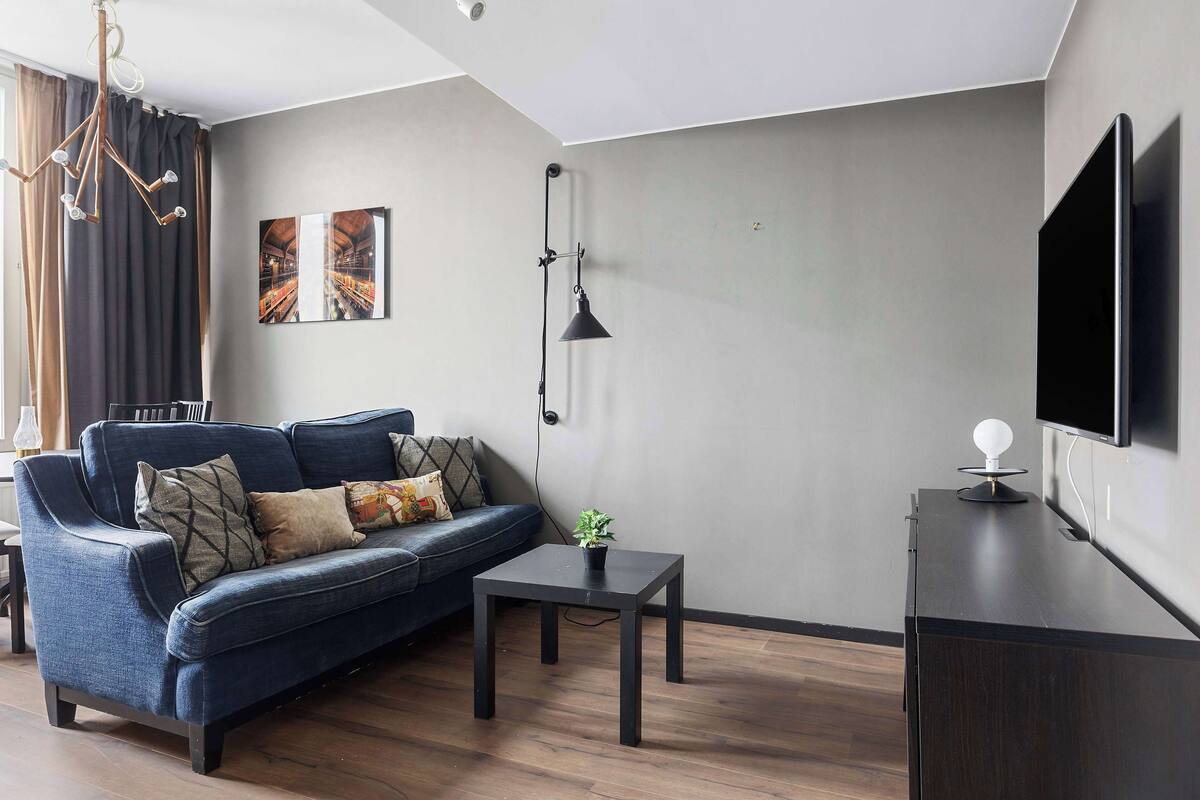 A modern living room with a dark blue sofa, a small table with a plant, a wall-mounted TV, and a ceiling light fixture.