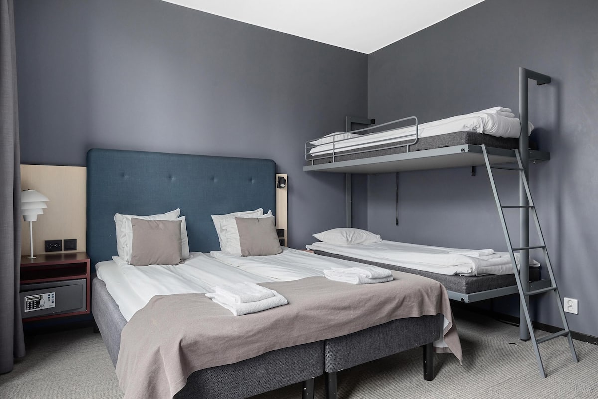 A room with a single bed, a bunk bed, a bedside table with a lamp, and a power outlet.