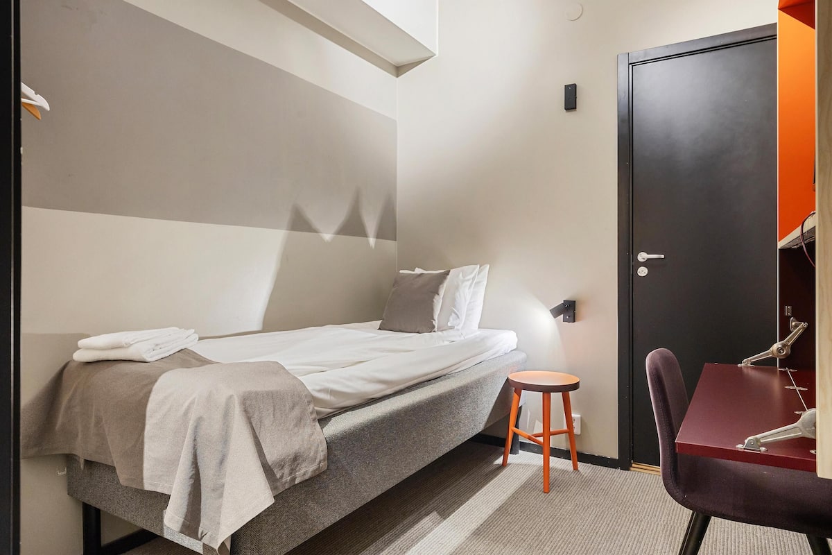 A compact hotel room with a bed, a small stool, a desk, and a wardrobe.
