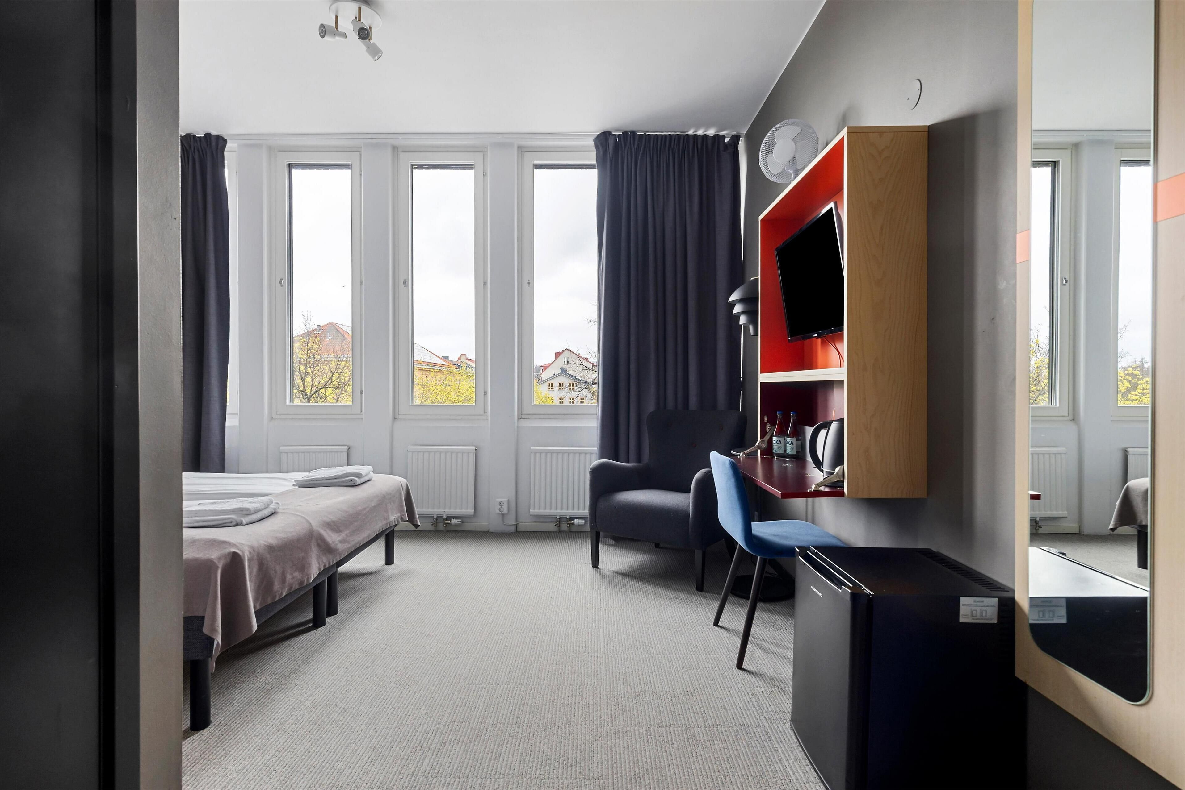A hotel room with a bed, a desk with a chair, a TV, and a view of buildings outside.