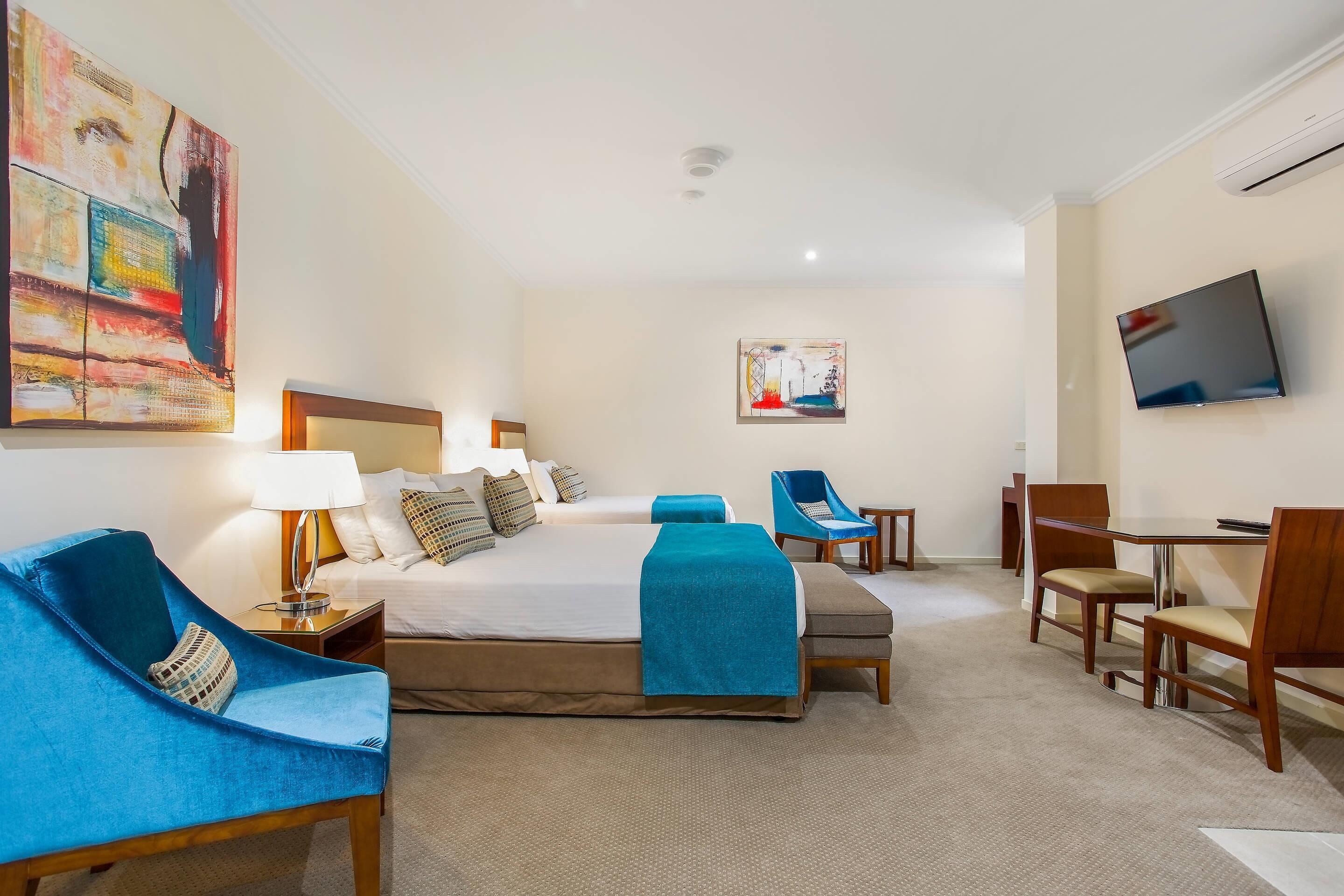 Habitación de hotel con una cama grande, dos mesitas de noche con lámparas, un escritorio con sillas, un televisor y obras de arte en las paredes.