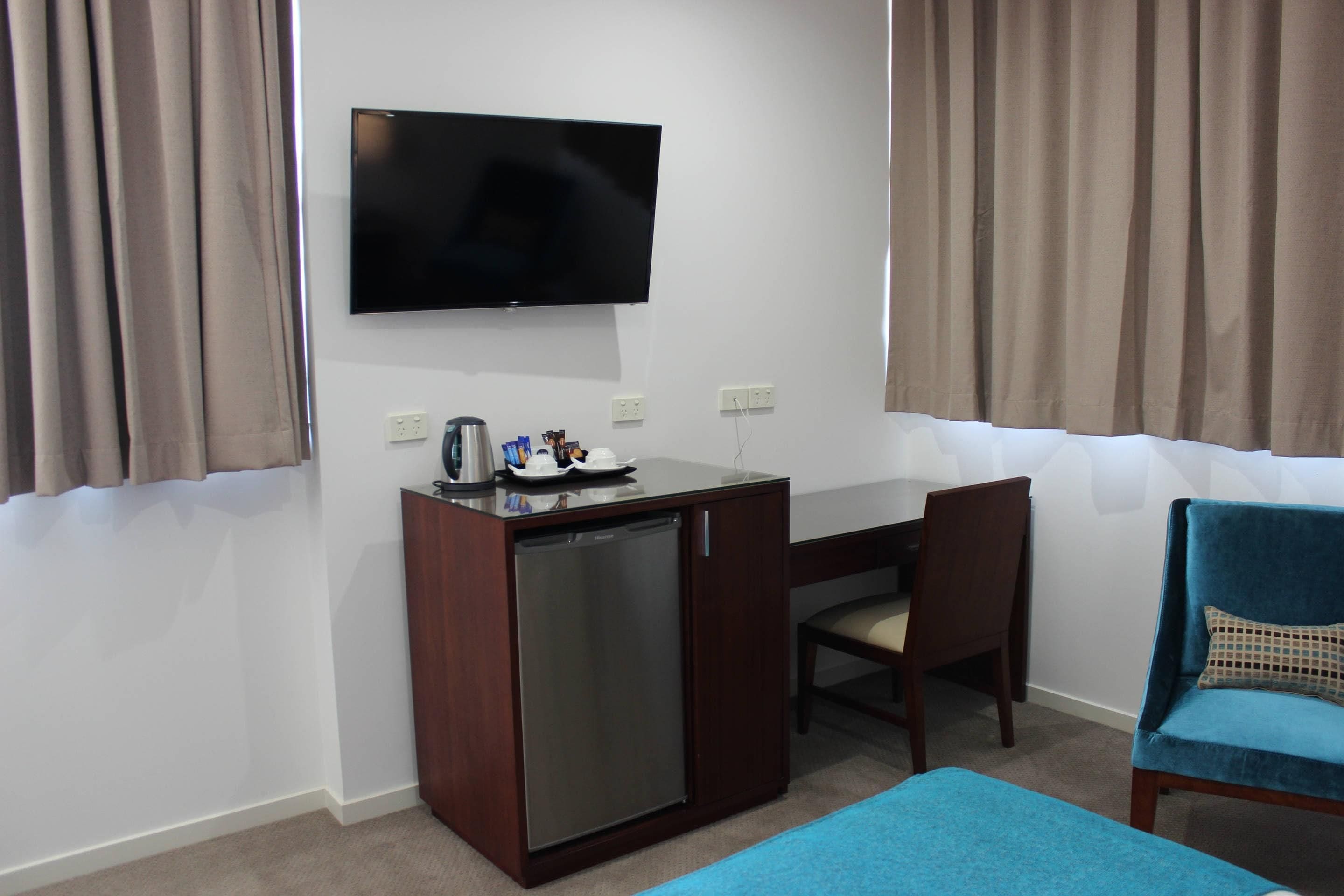 A compact hotel room with a flat-screen TV mounted on the wall, a small wooden desk with a refrigerator, a chair, and a blue sofa.