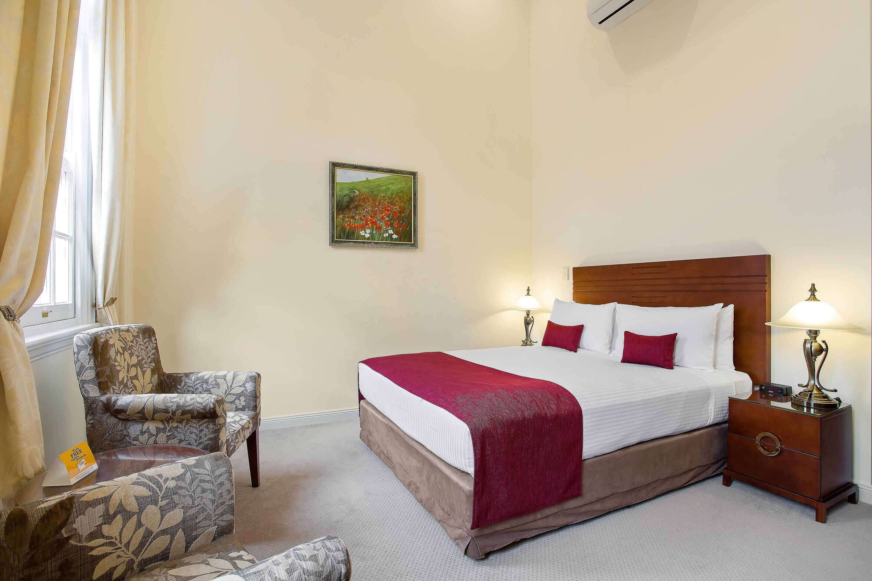 A hotel room with a bed, two bedside lamps, a chair, a nightstand, and a painting on the wall.