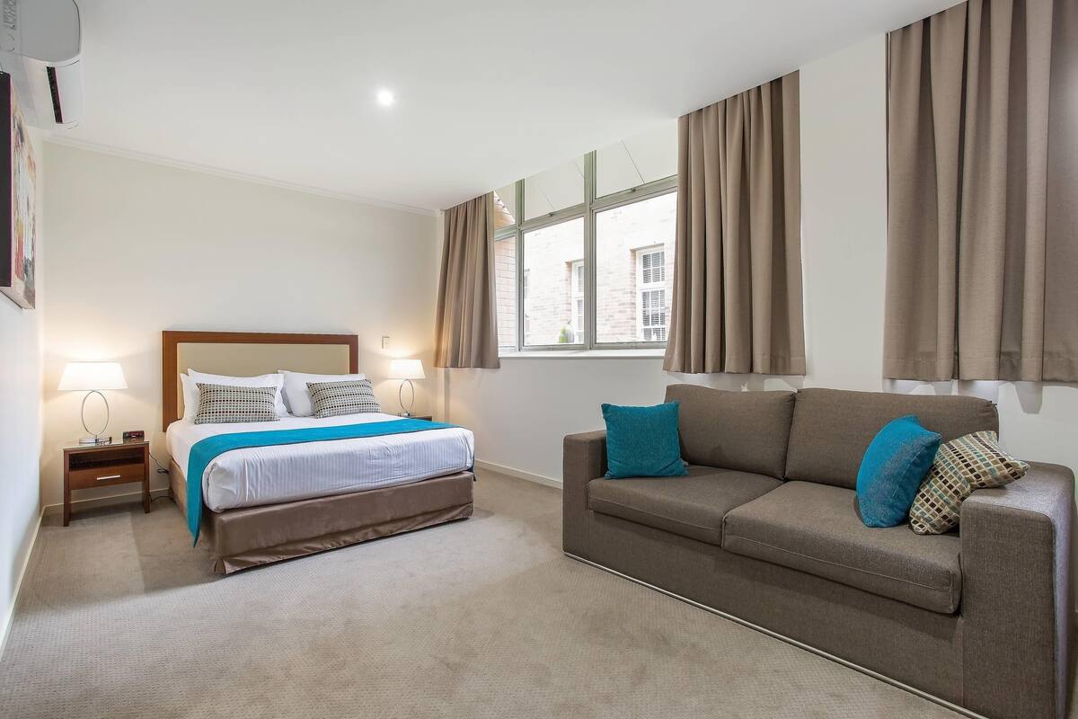 Ein Hotelzimmer mit einem Bett, einer Sitzecke, einem Nachttisch, einem Fenster mit Vorhängen und einer Wand-Klimaanlage.