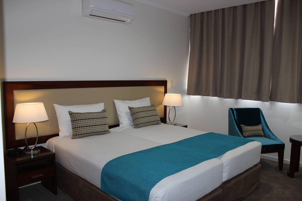 Ein Hotelzimmer mit einem großen Bett, zwei Nachttischlampen, einem Sessel und einem Fenster mit Vorhängen.