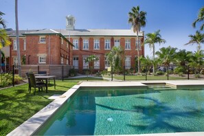 A swimming pool with a clear blue water, surrounded by a brick building with multiple windows and a fenced area with palm trees. - Quality Hotel Regent Rockhampton (Rockhampton)