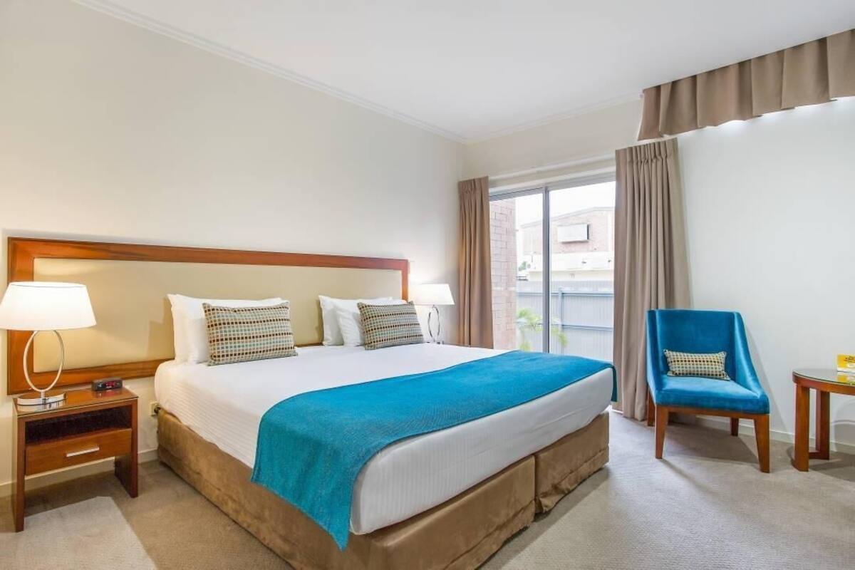Ein Hotelzimmer mit einem großen Bett, einem Nachttisch, einem Sessel und Blick ins Freie.