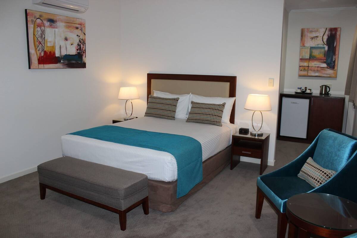 Ein Hotelzimmer mit einem Bett, zwei Nachttischen mit Lampen, einem Sessel, einem kleinen Tisch und Bildern an den Wänden.