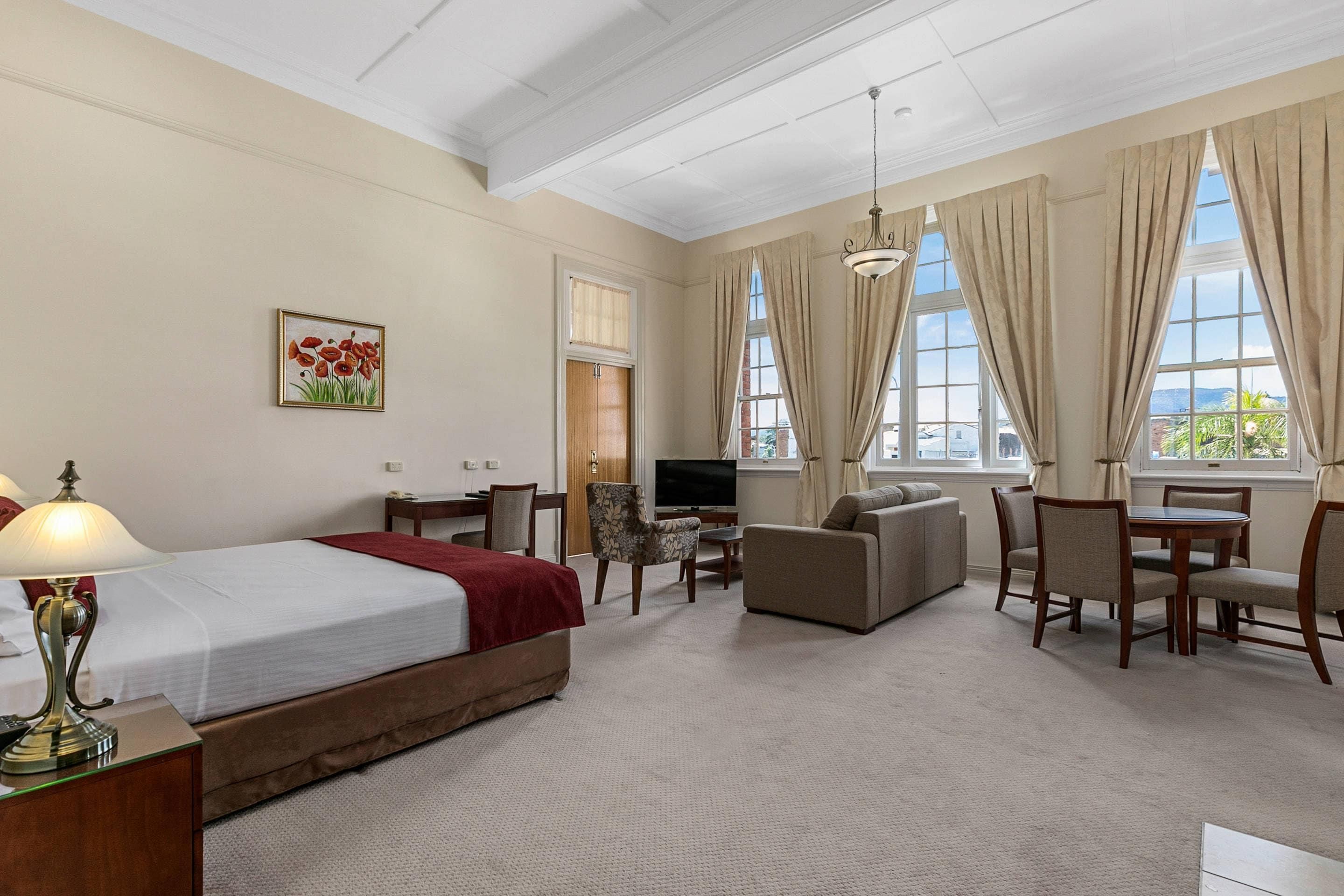 A hotel room with a bed, a desk with a lamp, a TV, a sofa, and a dining table with chairs.
