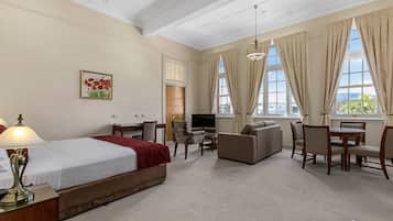 A hotel room with a bed, a desk with a lamp, a TV, a sofa, and a dining table with chairs.