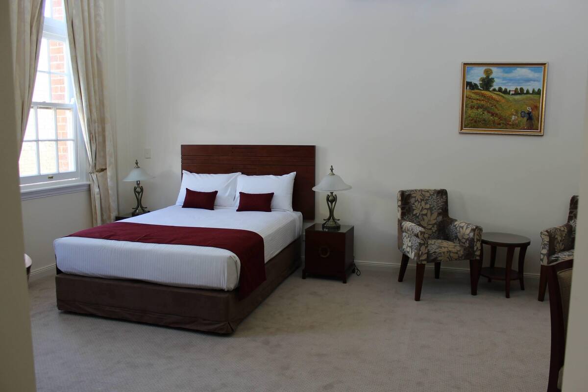 Ein Hotelzimmer mit einem großen Bett, zwei Nachttischen, einem Sessel, einem kleinen Tisch und einem Bild an der Wand.
