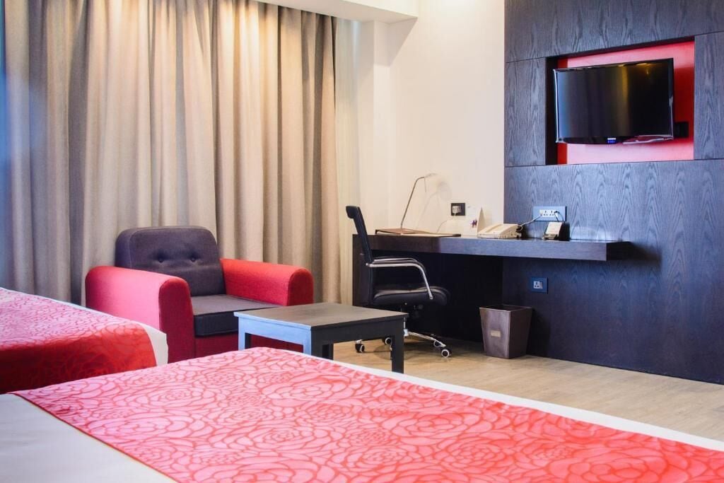 Habitación de hotel con cama, escritorio, silla, televisor montado en la pared y ventana con cortinas.