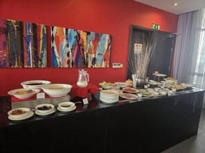 Mesa de buffet con diferentes alimentos y una jarra de leche, con pinturas abstractas coloridas en la pared y una puerta con letrero de salida de emergencia.
