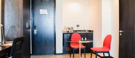 Una habitación compacta con una puerta de madera oscura, una pequeña cocina con lavamanos y encimera, una mesa de comedor con dos sillas rojas y una silla negra.
