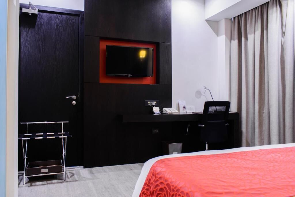 A hotel room with a bed, a desk with a computer, a television mounted on the wall, and a door.
