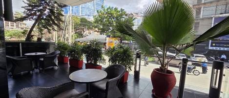 An outdoor seating area with wicker furniture and potted plants, overlooking a busy street with buildings and cars.