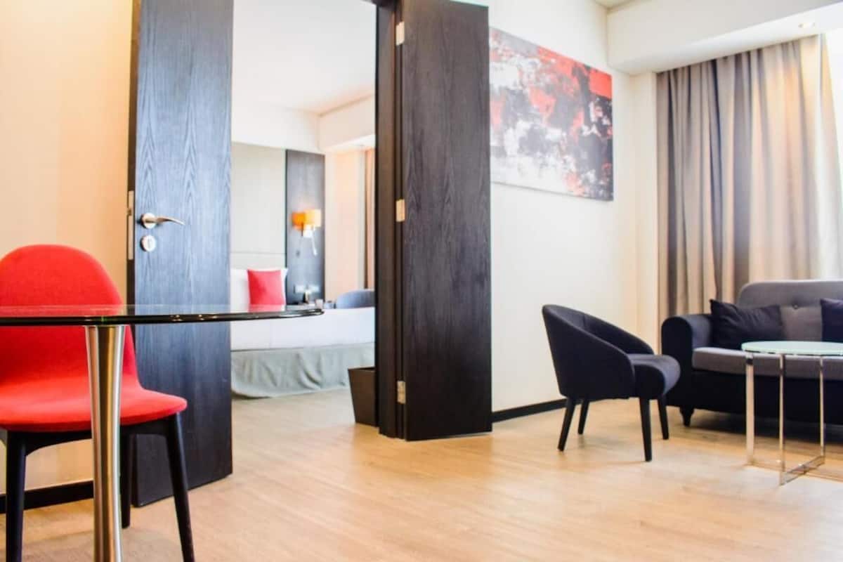 Habitación de hotel con cama, una silla roja, un sillón negro, una mesita y un ventanal grande con cortinas.