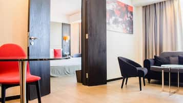 Quarto de hotel com uma cama, uma cadeira vermelha, uma poltrona preta, uma mesa pequena e uma janela grande com cortinas.