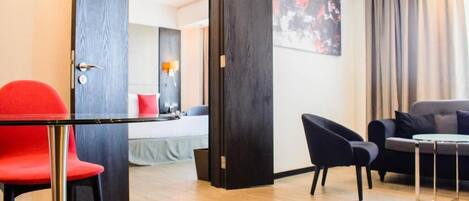A hotel room with a bed, a red chair, a black armchair, a small table, and a large window with curtains.