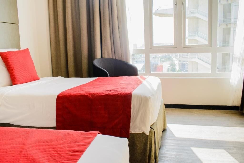 A hotel room with two beds, a view of the city through large windows, and a chair.