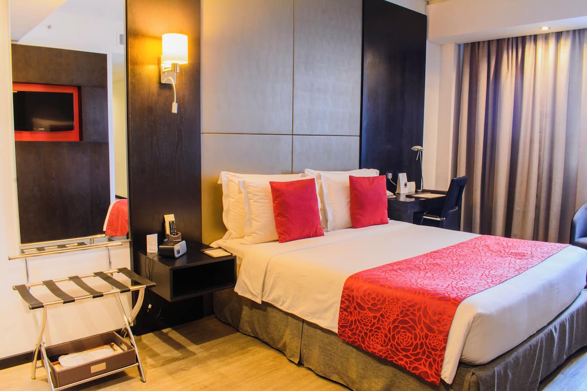 Habitación de hotel con una cama grande, un escritorio, una silla, una mesita de noche, una lámpara, un espejo y un televisor montado en la pared.