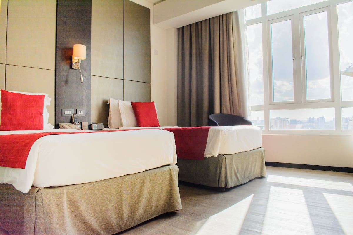 Habitación de hotel con dos camas, un escritorio, una silla, un ventanal y vistas a la ciudad.