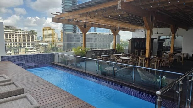 Una piscina en la azotea con terraza de madera y barandilla de vidrio, rodeada de mobiliario exterior y un área cubierta con vistas al horizonte de la ciudad.