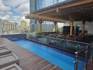 Una piscina en la azotea con terraza de madera y barandilla de vidrio, rodeada de mobiliario exterior y un área cubierta con vistas al horizonte de la ciudad.