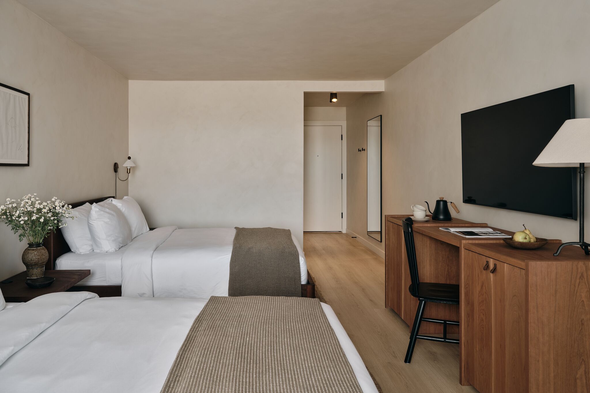 A hotel room with two beds, a desk, a chair, a television, and a door leading to another room.