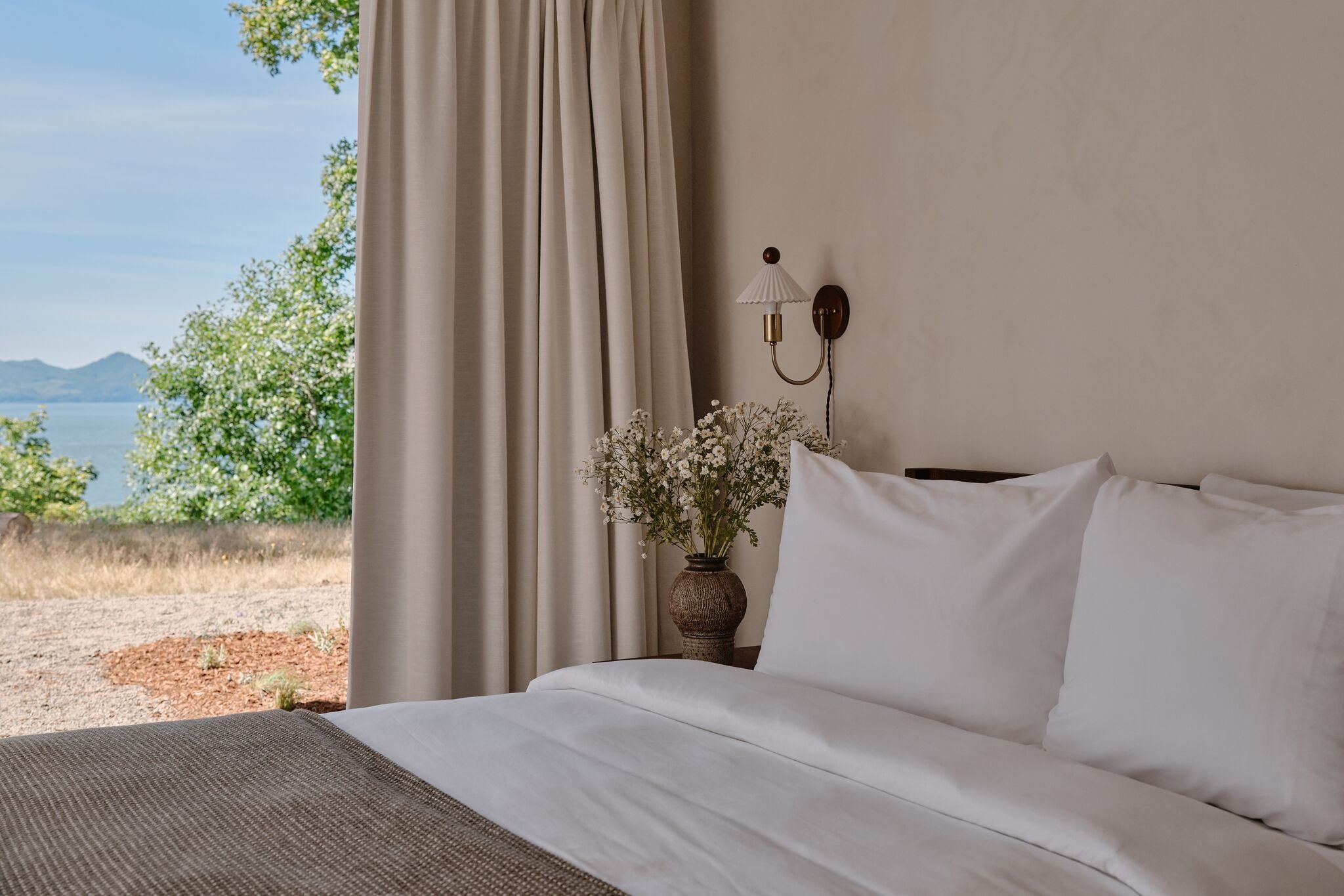 A bedroom with a bed, a vase with flowers, and a view of the outdoors.