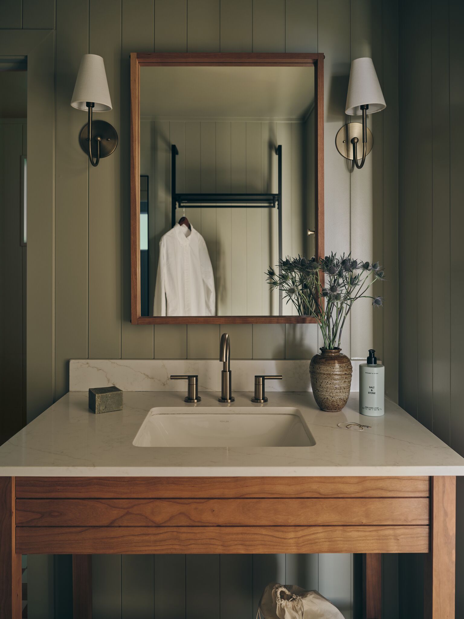 A bathroom with a wooden vanity, a large mirror, two wall-mounted lights, a sink with a modern faucet, a vase with dried flowers, and a bottle of hand soap.