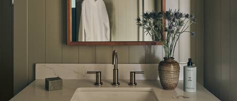 A bathroom with a wooden vanity, a large mirror, two wall-mounted lights, a sink with a modern faucet, a vase with dried flowers, and a bottle of hand soap.