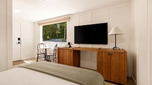 Une chambre à coucher avec un lit, un bureau avec une télévision, une chaise, une fenêtre donnant sur des arbres et une lampe.