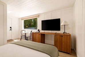 A bedroom with a bed, a desk with a TV, a chair, a window with a view of trees, and a lamp. - The Herondale Hotel (Astoria)