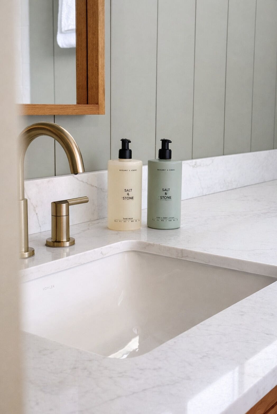 Un baño con un lavamanos blanco, un grifo dorado y dos dispensadores de jabón etiquetados 'Salt & Stone'.