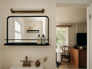 A bathroom with a mirror, a sink, a faucet, a vase with flowers, and a shelf with toiletries. - The Herondale Hotel (Astoria)