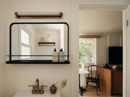 Un baño con espejo, lavamanos, grifo, un florero con flores y repisas con artículos de aseo.