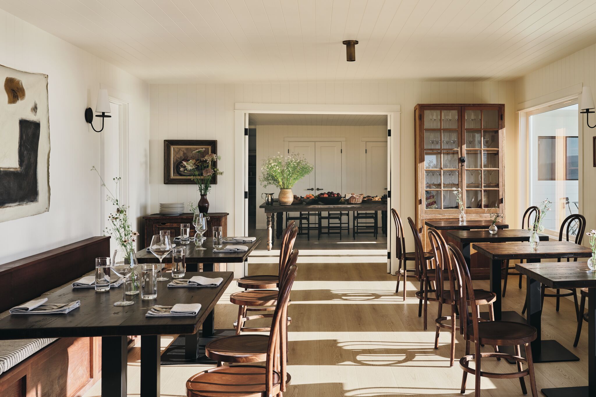 Un comedor con mesas y sillas de madera, una cocina al fondo y vistas por una puerta abierta.