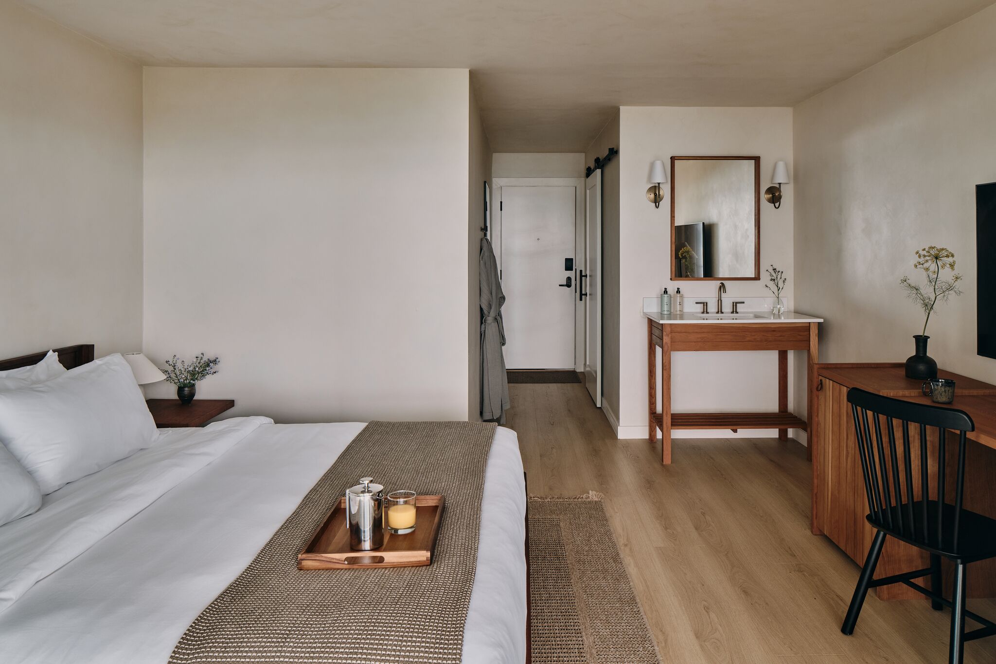 Hotelkamer met een bed, nachtkastje, een dienblad met koffiezetapparaat en kaars, een houten bureau met stoel, een badkamer met wastafel en spiegel, en een deur die naar een andere kamer leidt.