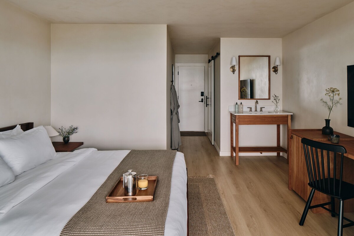 A hotel room with a bed, bedside table, a tray with a coffee pot and a candle, a wooden desk with a chair, a bathroom with a sink and mirror, and a door leading to another room.